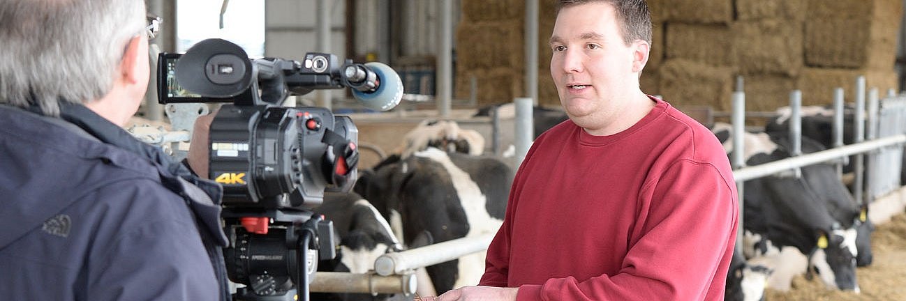 Das Foto zeigt einen Journalisten, der ein Videointerview mit einem Landwirt im Kuhstall führt. Sie stehen auf dem Futtertisch.