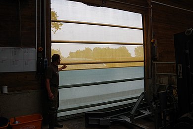 Ein Mann steht vor einem geschlossenen Windschutznetz am Stall. Klick führt zu Großansicht in neuem Fenster.
