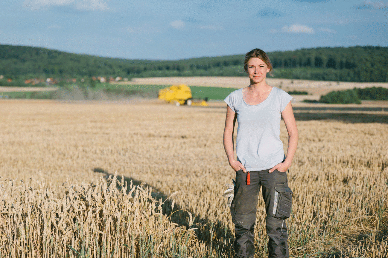 Eine Landwirtin steht in einem Weizenfeld und blickt in die Kamera. Klick führt zu Großansicht im neuen Fenster. 